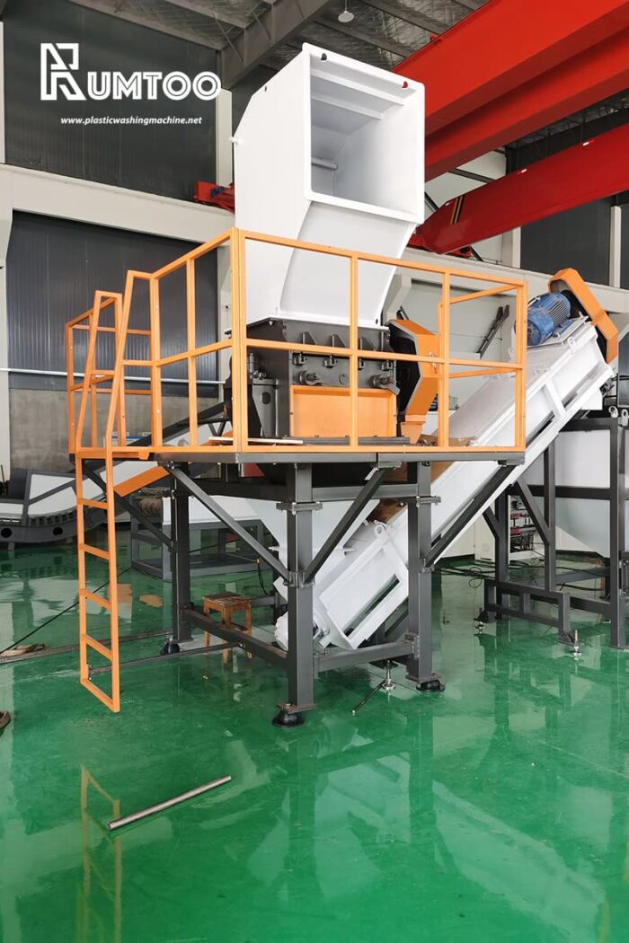 a large industrial machine, possibly a crusher or a shredder, used in a plastic recycling facility. The equipment is mounted on an elevated steel frame, painted orange, enhancing its visibility and safety. It features a substantial hopper at the top for feeding materials into the machine, which then leads to the processing unit housed within a white casing. Below the machine, a conveyor system appears to carry processed material away for further handling. The background of the image shows an industrial setting with other machinery and a green epoxy-coated floor, reflecting cleanliness and organization.