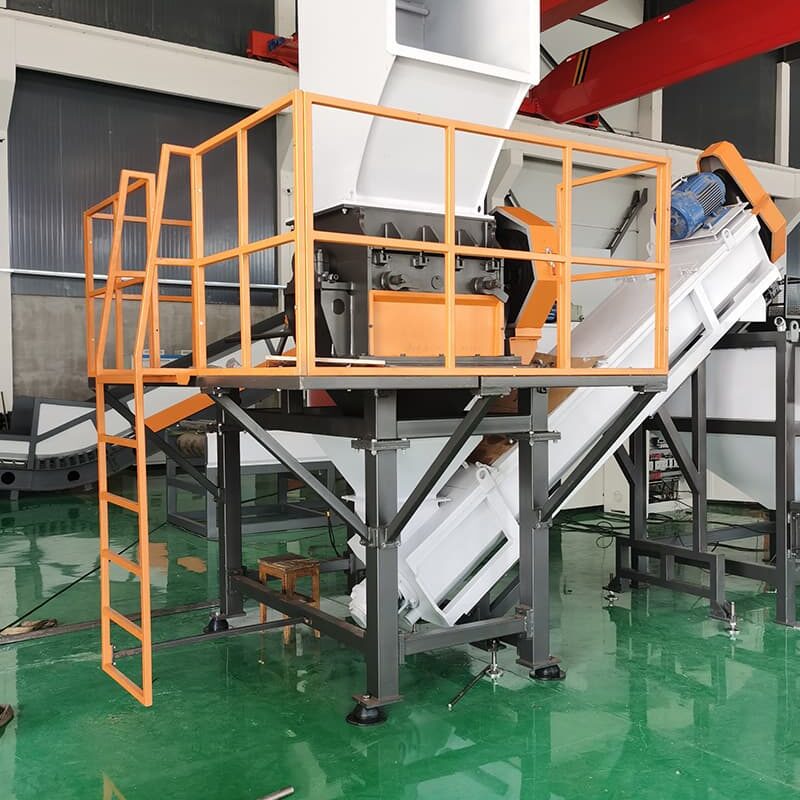 a large industrial machine, possibly a crusher or a shredder, used in a plastic recycling facility. The equipment is mounted on an elevated steel frame, painted orange, enhancing its visibility and safety. It features a substantial hopper at the top for feeding materials into the machine, which then leads to the processing unit housed within a white casing. Below the machine, a conveyor system appears to carry processed material away for further handling. The background of the image shows an industrial setting with other machinery and a green epoxy-coated floor, reflecting cleanliness and organization.