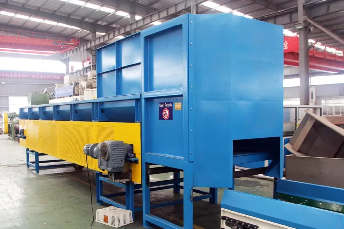 an Automatic Vertical Plastic Debaling Machine used in recycling facilities. This machine is designed to efficiently break down large bales of compacted plastic materials into looser forms that can be further processed and recycled. The debaling machine shown is robust, painted in blue and yellow for high visibility, and includes safety and operational signage. It features large blue housing that encloses the mechanical components necessary to separate the compacted materials, and a conveyor system integrated at the base to move the separated plastics to the next stage of the recycling process. This type of equipment is critical for handling and preparing bulk plastic waste for recycling, improving the efficiency of the recycling workflow.