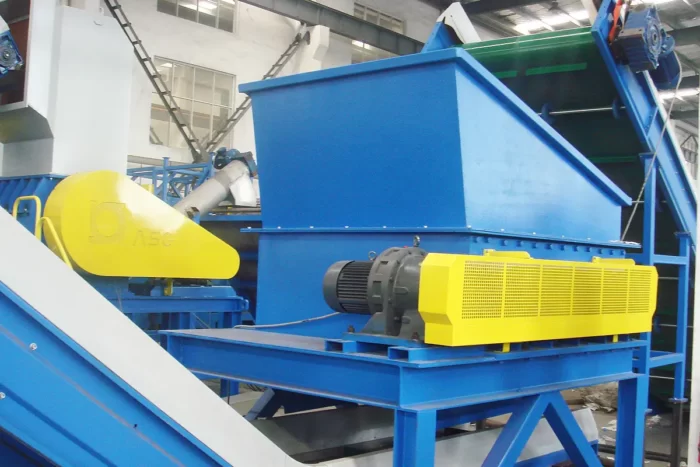 a horizontal debaler machine, primarily used in recycling facilities to break down and separate materials from baled plastic or paper products. The machine is robust, featuring a predominantly blue and yellow color scheme, which highlights its industrial nature. A large yellow motor and conveyor system indicate where the bales are processed and moved through the machine. The setup also includes protective housing and structural supports in blue, ensuring safety and durability. This machinery is essential for initial processing in recycling operations, preparing materials for further shredding or washing.