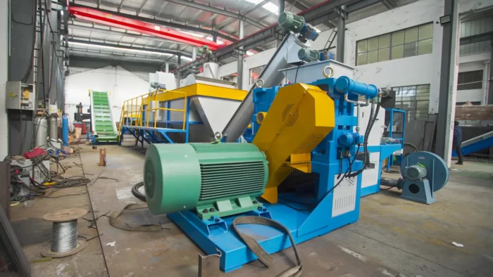 a comprehensive view of an industrial plastic recycling facility, featuring a variety of specialized equipment. This setup includes a series of machines such as conveyors, shredders, and separators, all crucial for the recycling process. The machinery is predominantly painted in vibrant blue and yellow, colors often used to denote machinery and caution in industrial environments. This particular section of the facility appears to focus on the initial stages of recycling where plastics are sorted, cleaned, and prepared for further processing. Visible in the scene are large motors and mechanical parts, suggesting these machines handle heavy-duty processing tasks like shredding and crushing. The background shows additional parts of the facility, hinting at a large-scale operation designed to process significant volumes of material efficiently.