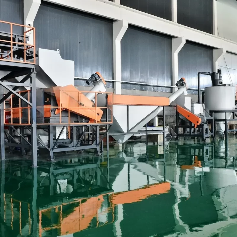 depicts a sophisticated plastic recycling facility equipped with LDPE (Low-Density Polyethylene) recycling equipment. The setup features various machines including conveyors, crushers, and wash stations that are strategically placed over a large water tank for the separation and washing of recycled materials. The system's framework is painted in a mix of gray and orange, providing a stark contrast to the industrial background. This type of facility is typically used for the recycling of soft plastics, such as plastic bags and films, which undergo processes including shredding, washing, and drying to prepare them for reintegration into the production cycle. The presence of the water tank is indicative of the washing process, where plastics are cleansed of impurities such as soil, labels, and adhesives. Facilities like this play a crucial role in minimizing waste and promoting sustainable practices within the plastics industry.