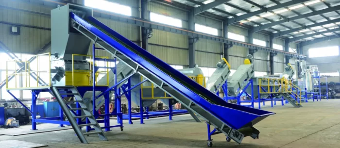 an industrial facility for recycling rigid plastics. The system includes various machines and conveyors arranged in sequence, likely designed for the shredding, washing, sorting, and reprocessing of plastic materials. The equipment is predominantly blue and yellow, enhancing visibility and safety in the industrial environment. This type of recycling line is crucial for processing large volumes of plastic waste, converting it into reusable raw materials, thereby supporting sustainable practices and reducing environmental impact. Such systems are integral to modern waste management and recycling operations.