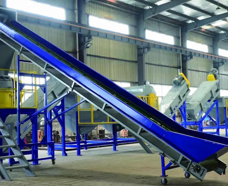 an industrial facility for recycling rigid plastics. The system includes various machines and conveyors arranged in sequence, likely designed for the shredding, washing, sorting, and reprocessing of plastic materials. The equipment is predominantly blue and yellow, enhancing visibility and safety in the industrial environment. This type of recycling line is crucial for processing large volumes of plastic waste, converting it into reusable raw materials, thereby supporting sustainable practices and reducing environmental impact. Such systems are integral to modern waste management and recycling operations.