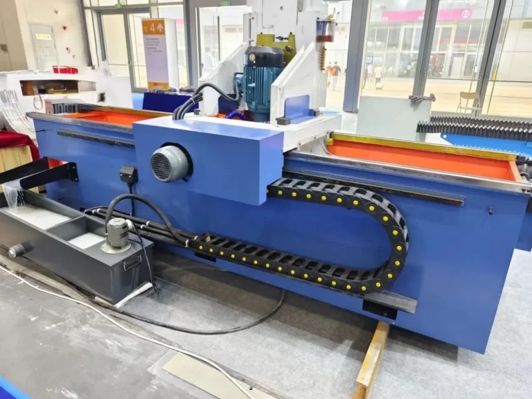 an Auto Knife Grinder, a specialized machine used primarily for sharpening the blades used in industrial cutting or processing machinery. This particular model features a long, horizontal grinding bed where knives or blades are secured and sharpened, ensuring they maintain optimal cutting efficiency. The machine is painted in a vibrant blue with red and orange accents, typical for industrial equipment to enhance visibility and safety.
