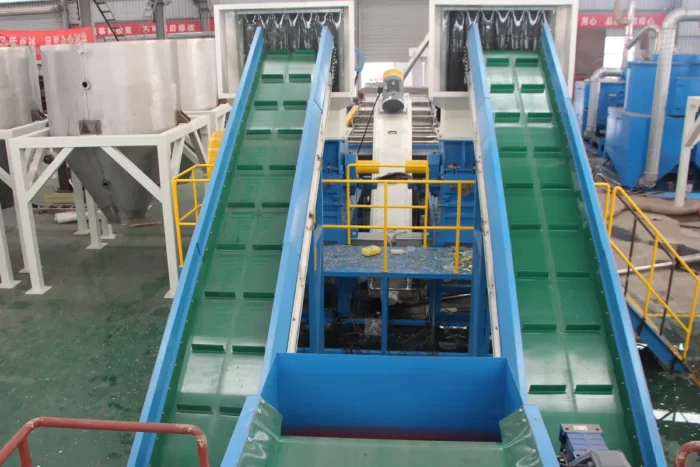 an operational scene within a plastic recycling facility, focusing on a series of conveyor belts used for transporting plastic materials through various stages of the recycling process. These conveyors are part of a larger system that likely includes sorting, cleaning, and shredding operations. The equipment is painted in vivid green and blue, enhancing visibility and safety within the industrial setting. In the background, there are various large tanks and machinery, possibly for washing or further processing the plastics. The setup shown is typical for facilities handling large volumes of recyclable materials, emphasizing efficient flow and processing to convert waste into reusable resources. Such facilities play a crucial role in environmental sustainability by reducing landfill use and providing recycled materials for new products.