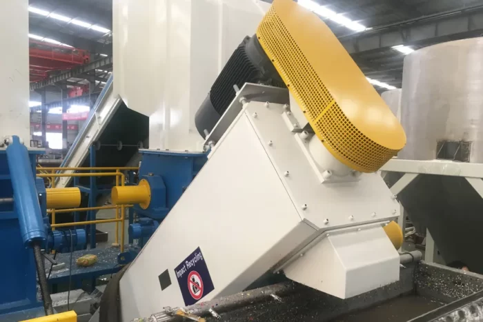 an industrial friction screw washer, a machine primarily used in the recycling process to clean plastic flake. This equipment works by using mechanical friction and water to remove contaminants like dirt, labels, and adhesives from plastic materials. The friction screw washer in the image features a robust design, with a large inclined screw enclosed in a housing. The housing is primarily white, with a yellow protective cover over the motor, enhancing safety and visibility in an industrial setting. Such washers are essential components in recycling facilities, ensuring that plastic flakes are thoroughly cleaned before they proceed to the next stages of recycling, such as melting and pelletizing.