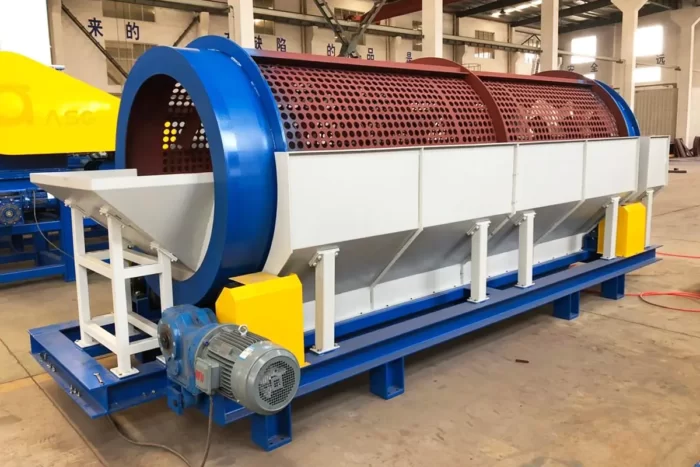 a large, industrial trommel screen, a vital piece of equipment commonly used in recycling facilities to separate materials by size. This cylindrical screen rotates to sort materials, with smaller particles falling through the holes while larger items continue to the end of the trommel.